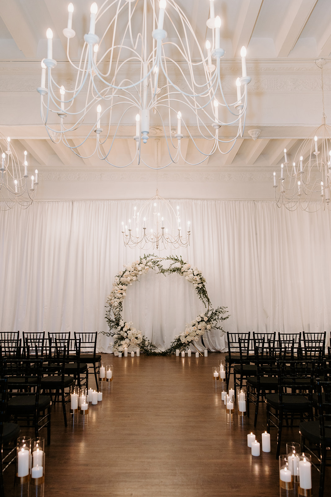 Arbor adorned with White Flowers in the ceremony room with black chairs and white candles on the floor.