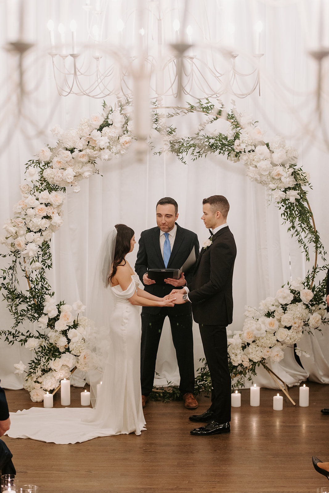 Bride and groom giving their vows with their officiant under a wedding arbor adorned with white flowers.